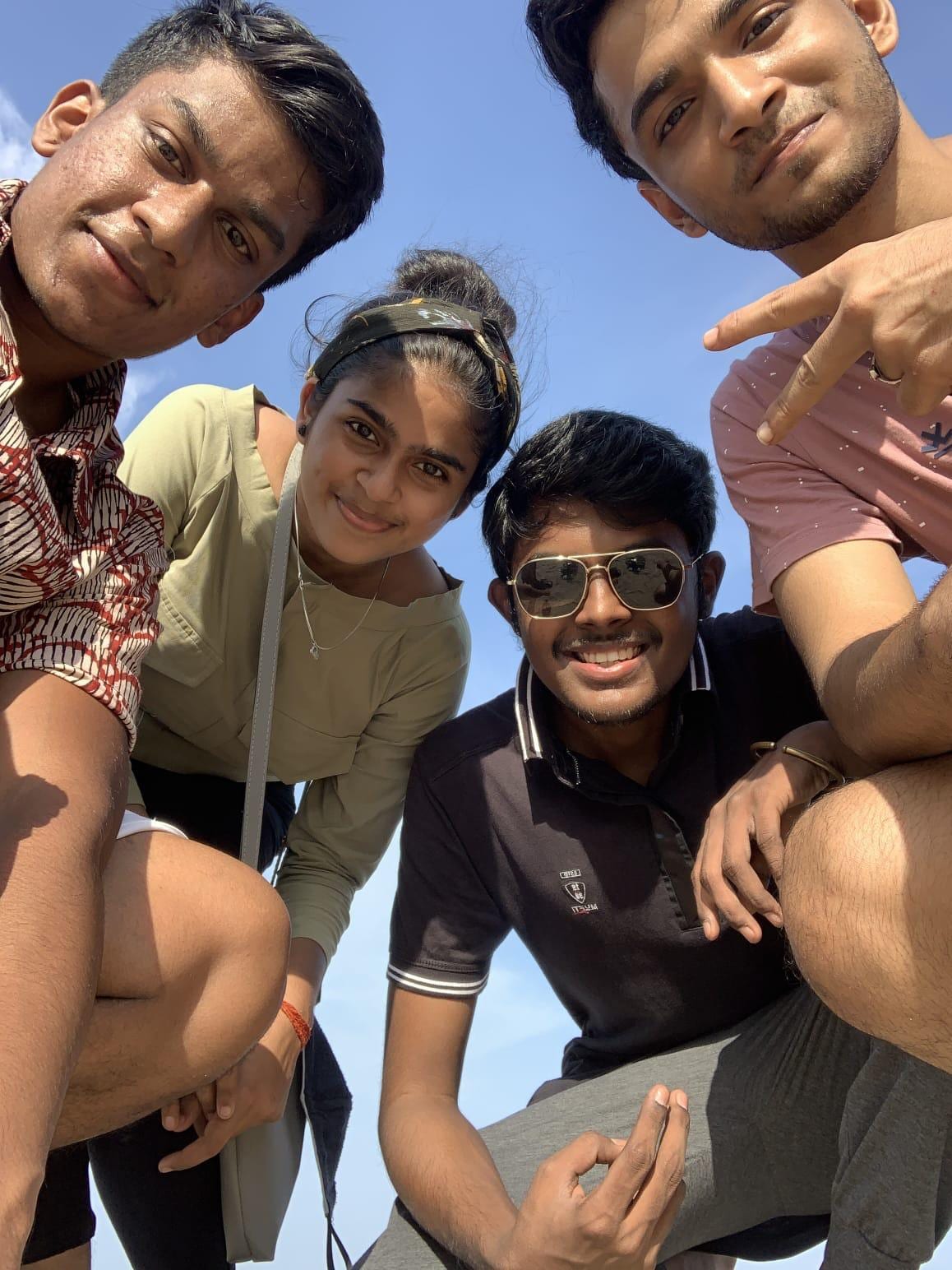 Thilak and three friends crouched shoulder to shoulder, photographed from below against a bright blue sky, one of them holding up a peace sign.