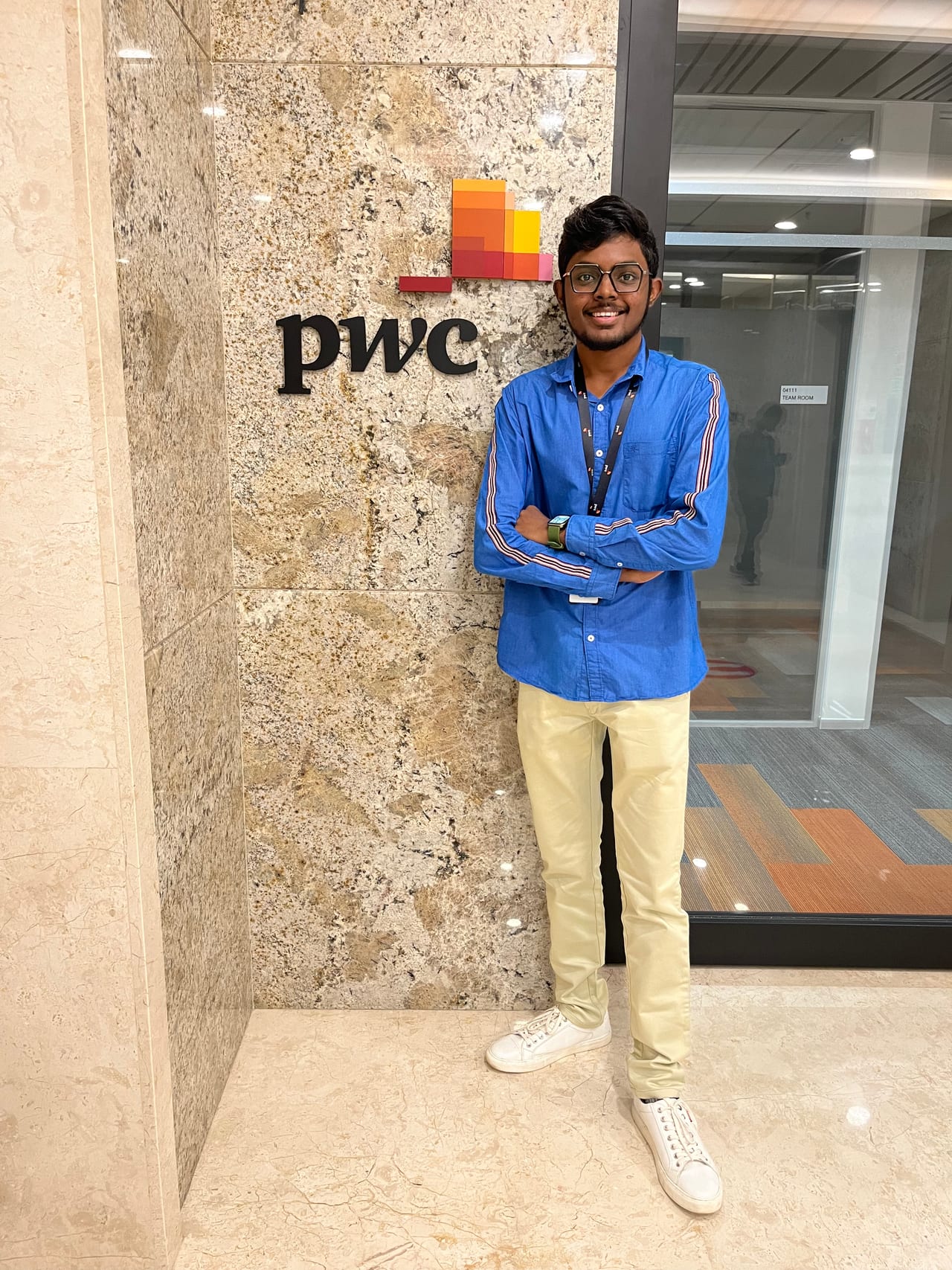 Thilak with his arms folded beside the PwC sign in an office lobby, wearing a company lanyard.