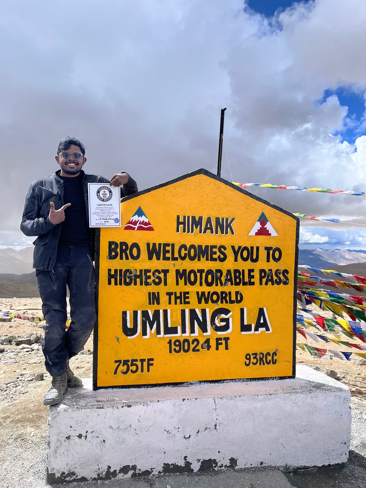 Thilak in motorcycle gear beside the Umling La marker, a yellow sign reading highest motorable pass in the world at 19,024 feet, with prayer flags and bare mountains behind.