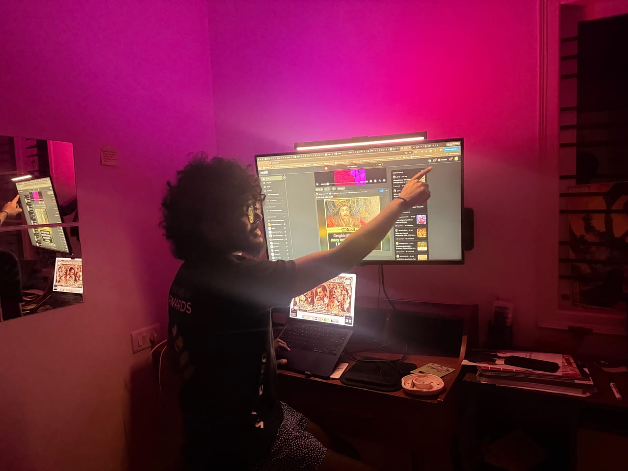 Thilak pointing at a large wall mounted monitor covered in open browser tabs, in a room lit magenta, a laptop open on the desk below.